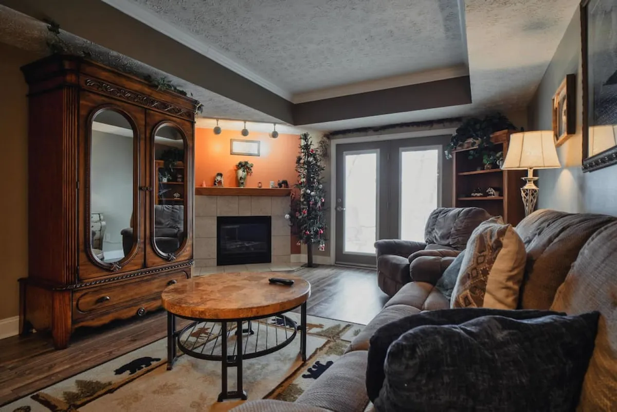 A spacious living room boasting overstuffed sofas, a coffee table, a wooden armoire, and a glowing fireplace beneath large windows. The atmosphere is both cozy and open, ideal for socializing or unwinding by the fire.