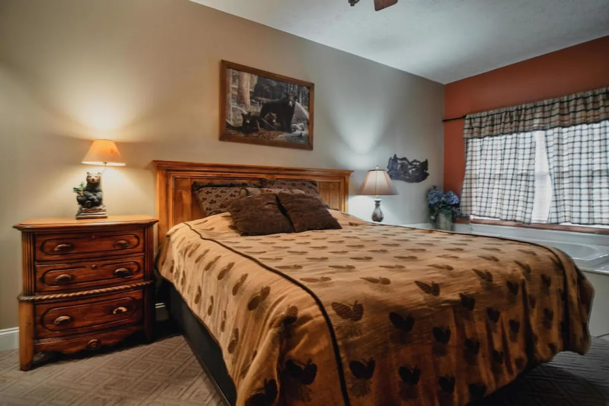 The image shows a warm, inviting bedroom with a plush bed adorned in a patterned blanket, a soft-glowing nightstand lamp, framed art on the wall, and a window dressed in cozy checkered curtains letting in gentle natural light.
