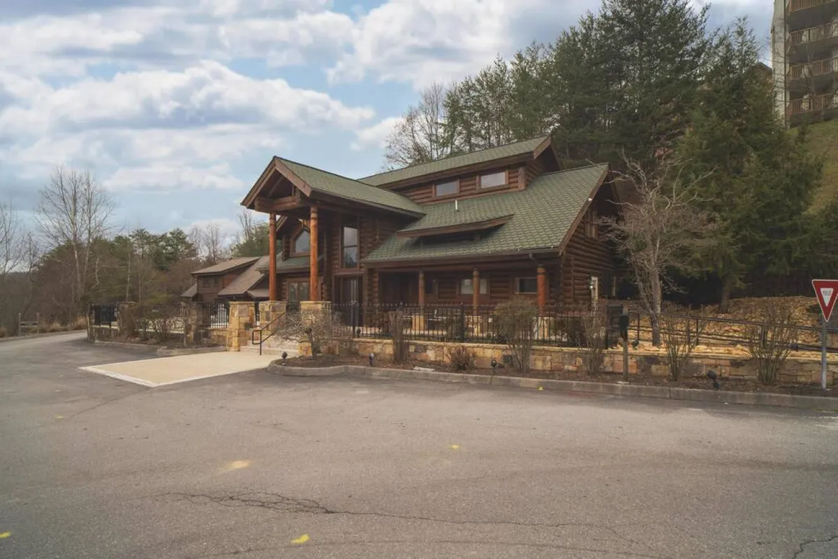 The photo features a cozy log cabin structure with a green roof, stone foundation, and a landscaped garden. The inviting porch hints at outdoor gatherings, grilling, and moments shared after a day of adventure. The scene is vibrant and welcoming, with the surrounding greenery suggesting connections to both nature and group activities.