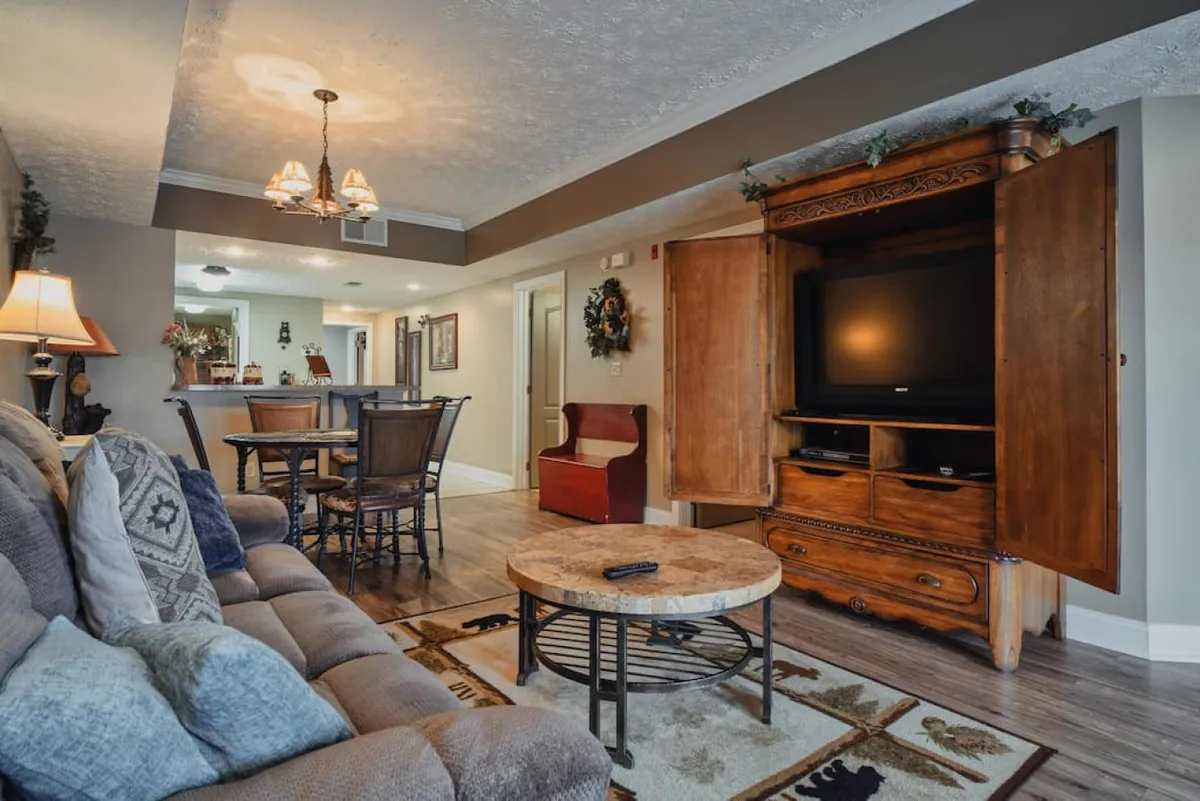 A warmly lit living area featuring a plush sofa with decorative pillows, a dining table and chairs set for shared meals, and an ornate wooden entertainment unit. The space invites relaxation and togetherness, with hints of family gatherings and cozy evenings.