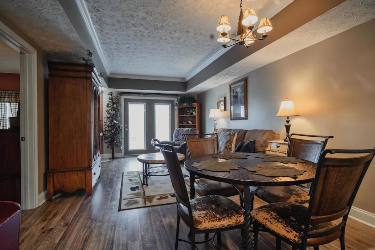 A spacious wooden dining table sits beside a comfortable seating area, framed by large windows that let in abundant natural light. Decorative lighting and artwork enhance the warm, welcoming atmosphere, and the space is clearly designed for both large feasts and intimate dinners.