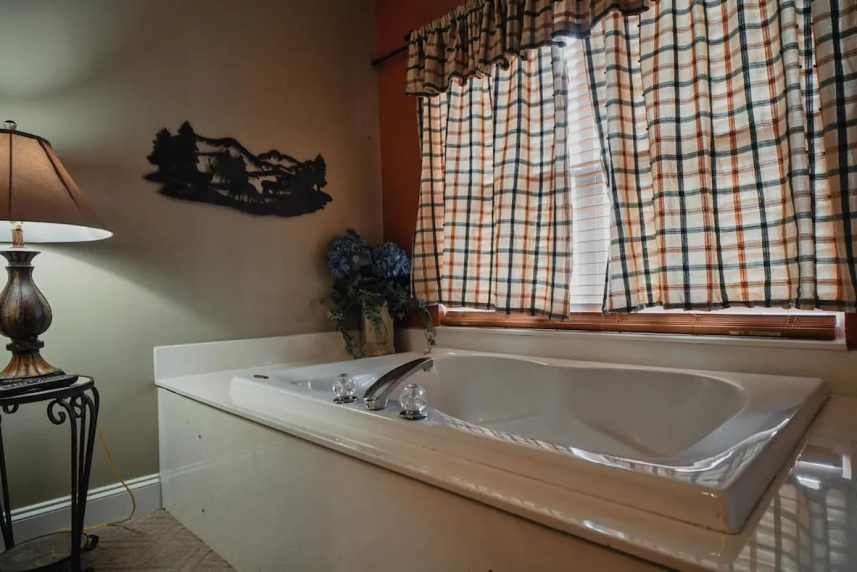 A tranquil bathroom scene with a standalone bathtub, elegant lamp, flowing curtains, decorative wall art, and fresh flowers. The mood is spa-like and serene, perfect for relaxation and self-care.