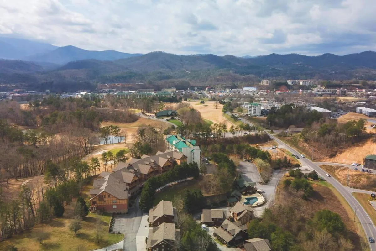 A broad exterior view featuring lodge-style resort buildings set against a dramatic mountain range, with a golf course and a hint of a charming town area nearby. The scene feels inviting, adventurous, and connected to the local culture.